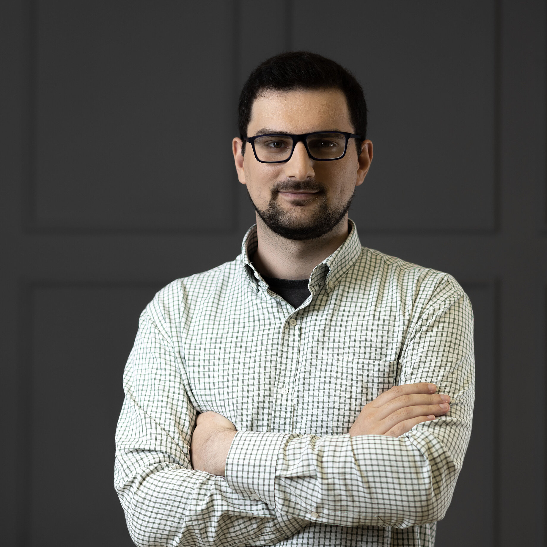 A man standing straight with his arms crossed in front of a dark and patterned background. He has dark short hair and has a trimmed beared. He is also wearing glasses and a light and patterend shirt.