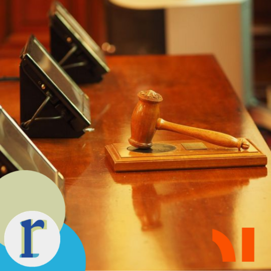 A wooden gavel resting on its sound block on a polished wooden table, with microphone stands and name-plate holders lined up beside it, in what looks like a committee or courtroom setting.