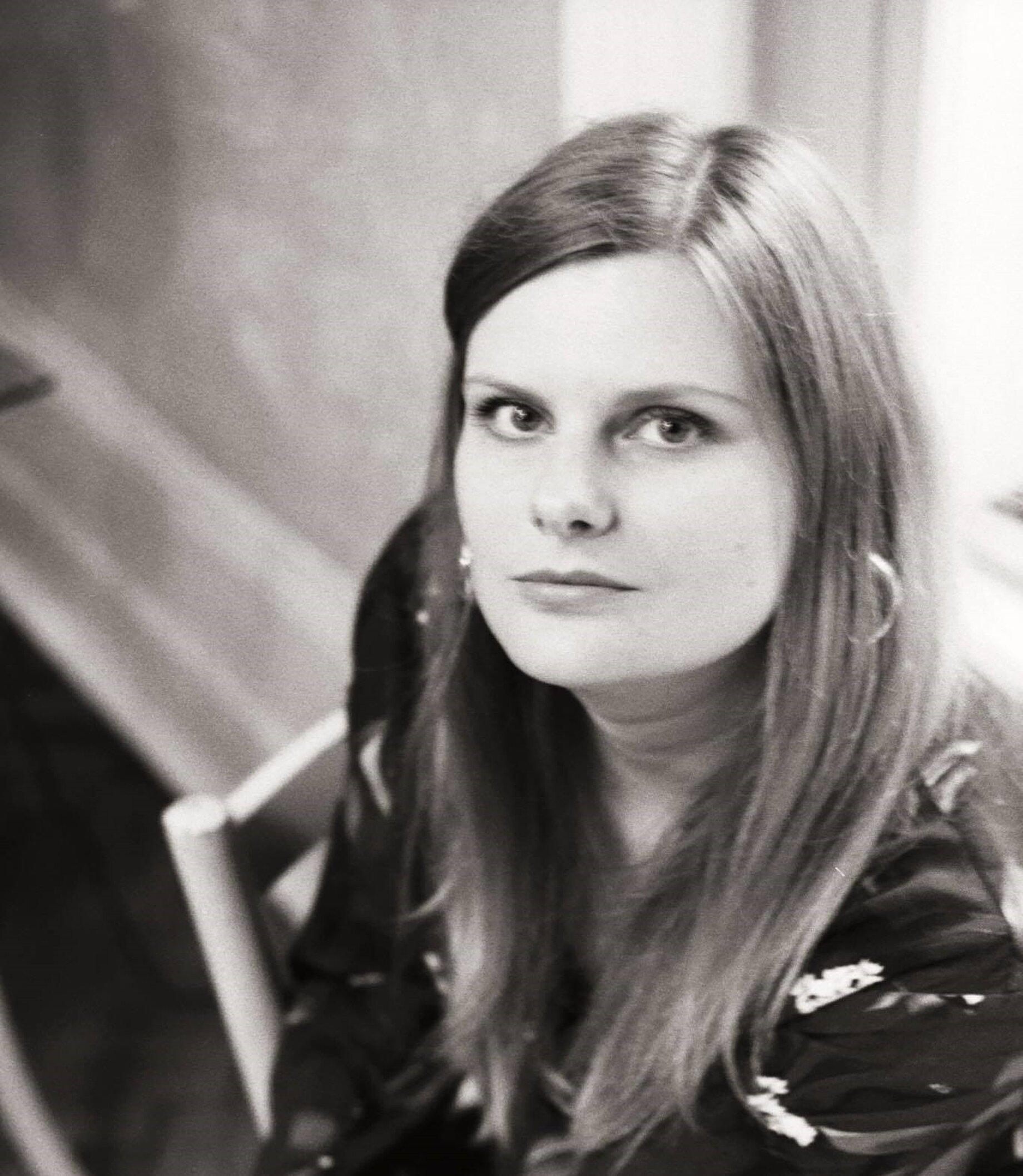 A woman in a black and white picture with long and straight hair. She is wearing earrings and is sitting on a chair