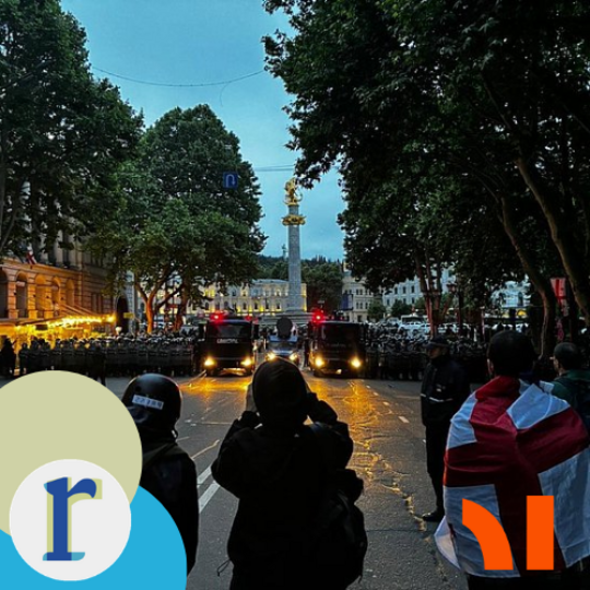 An evening street scene with riot police and armoured vehicles blocking a tree-lined avenue near a lit monument topped with a golden statue, with bystanders and a red-and-white flag in the foreground, apparently in Tbilisi, Georgia.