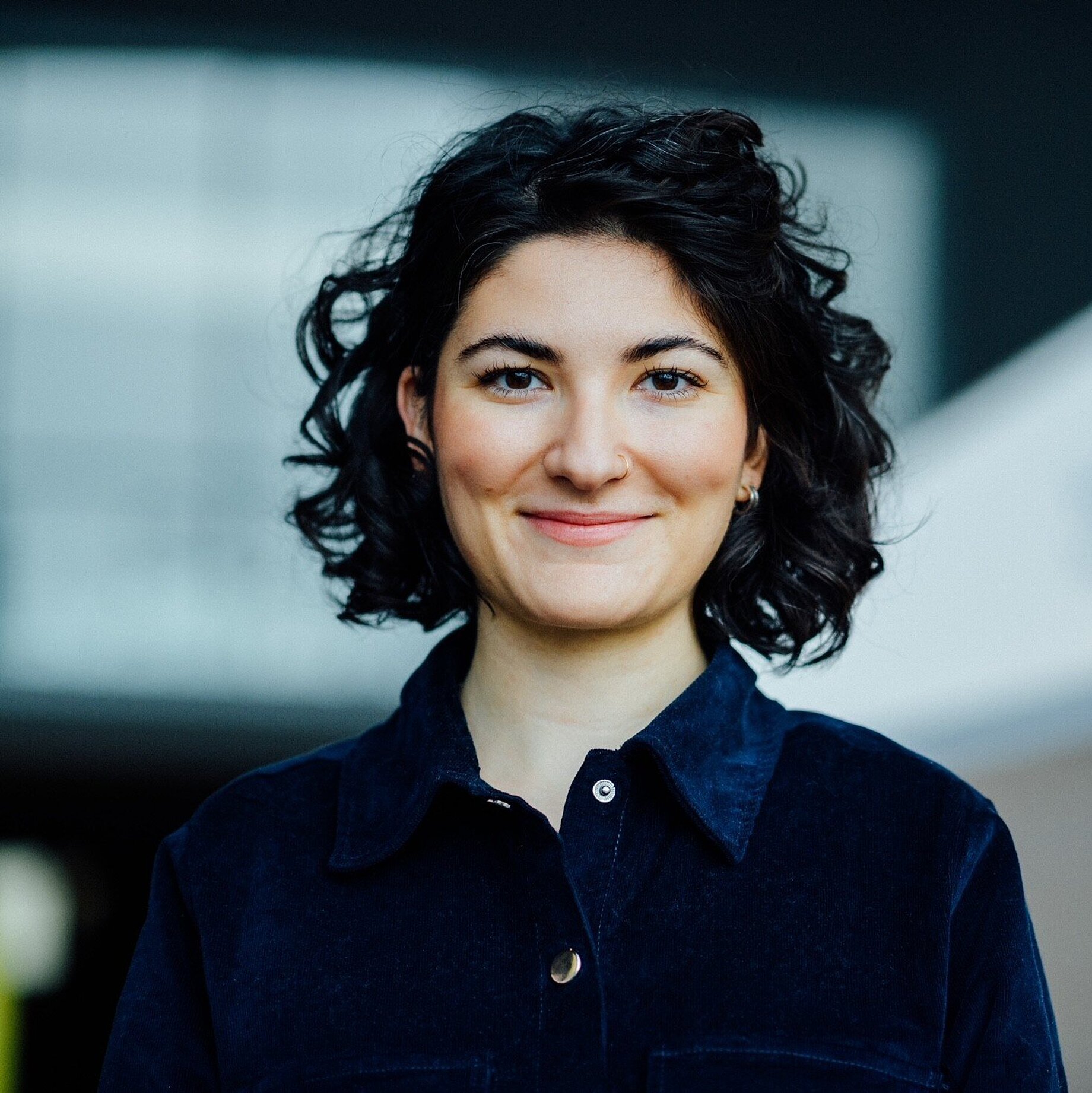 A woman with short wavy black hair who is wearing a nose and earring and a denim jacket.