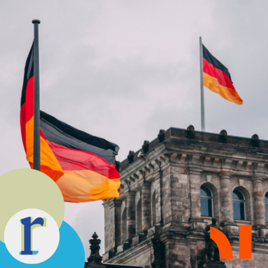Two German flags flying on poles in front of the Reichstag building's stone tower, under an overcast sky.
