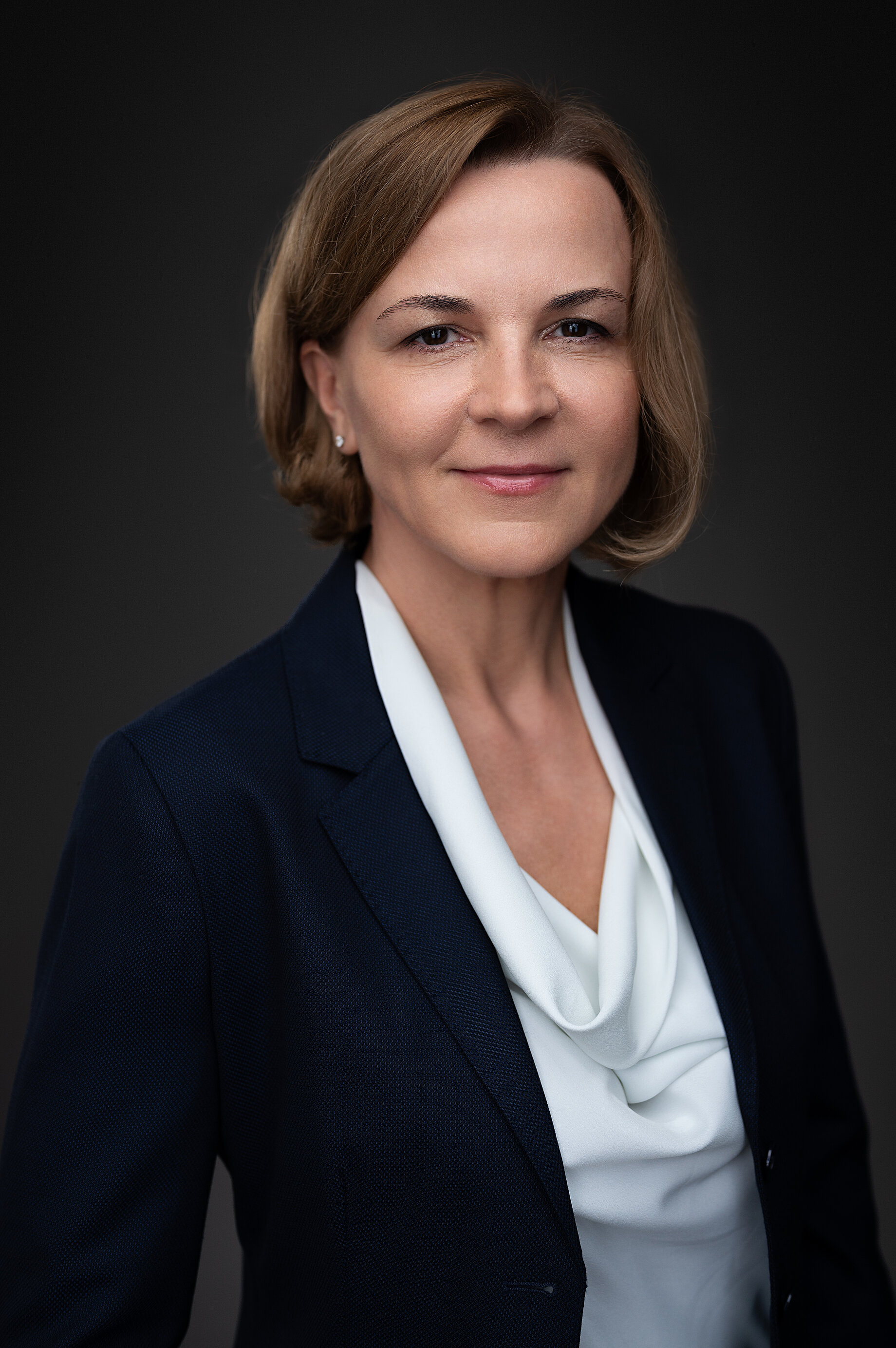A woman with short light brown hair in wearing a navy blue suit and diamond earrings