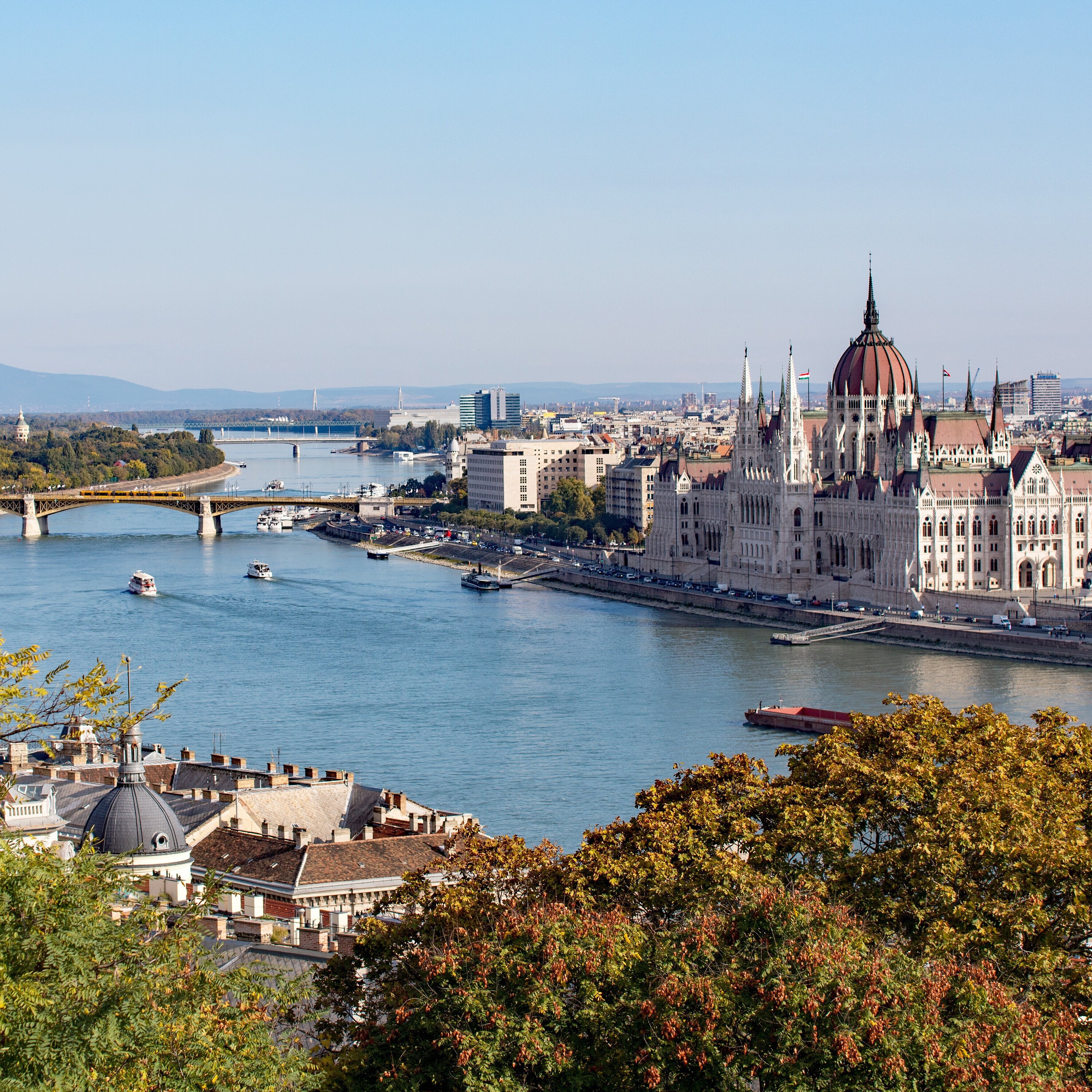 Photo of the Hungarian Parliament building taken from a hill across the Danube river. It shows trees in the front, the Danube and the Parliament in the background.