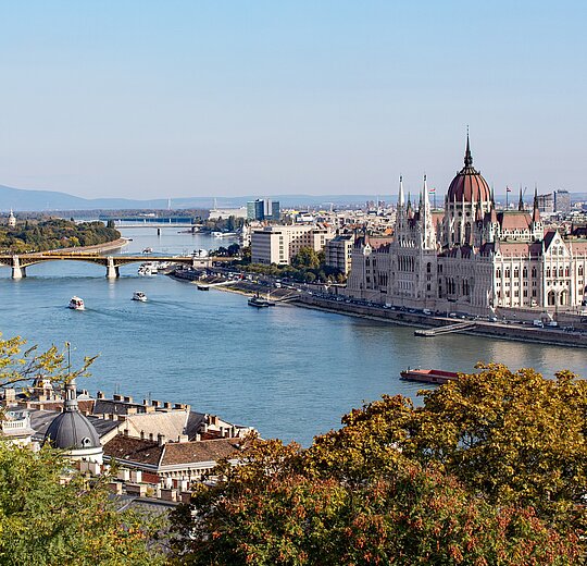 Photo of the Hungarian Parliament building taken from a hill across the Danube river. It shows trees in the front, the Danube and the Parliament in the background.