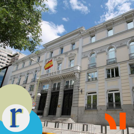 A tan stone building facade with the Spanish flag hanging above an arched entrance and temporary metal barriers set up outside, in an urban street setting.