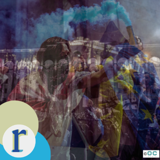 A blurred, layered image of a nighttime street protest, with a person holding up a blue smoke flare, an EU flag and Goergian flag, and a line of riot police or barriers in the background (photo credit: Mariam Nikuradze / OC Media).