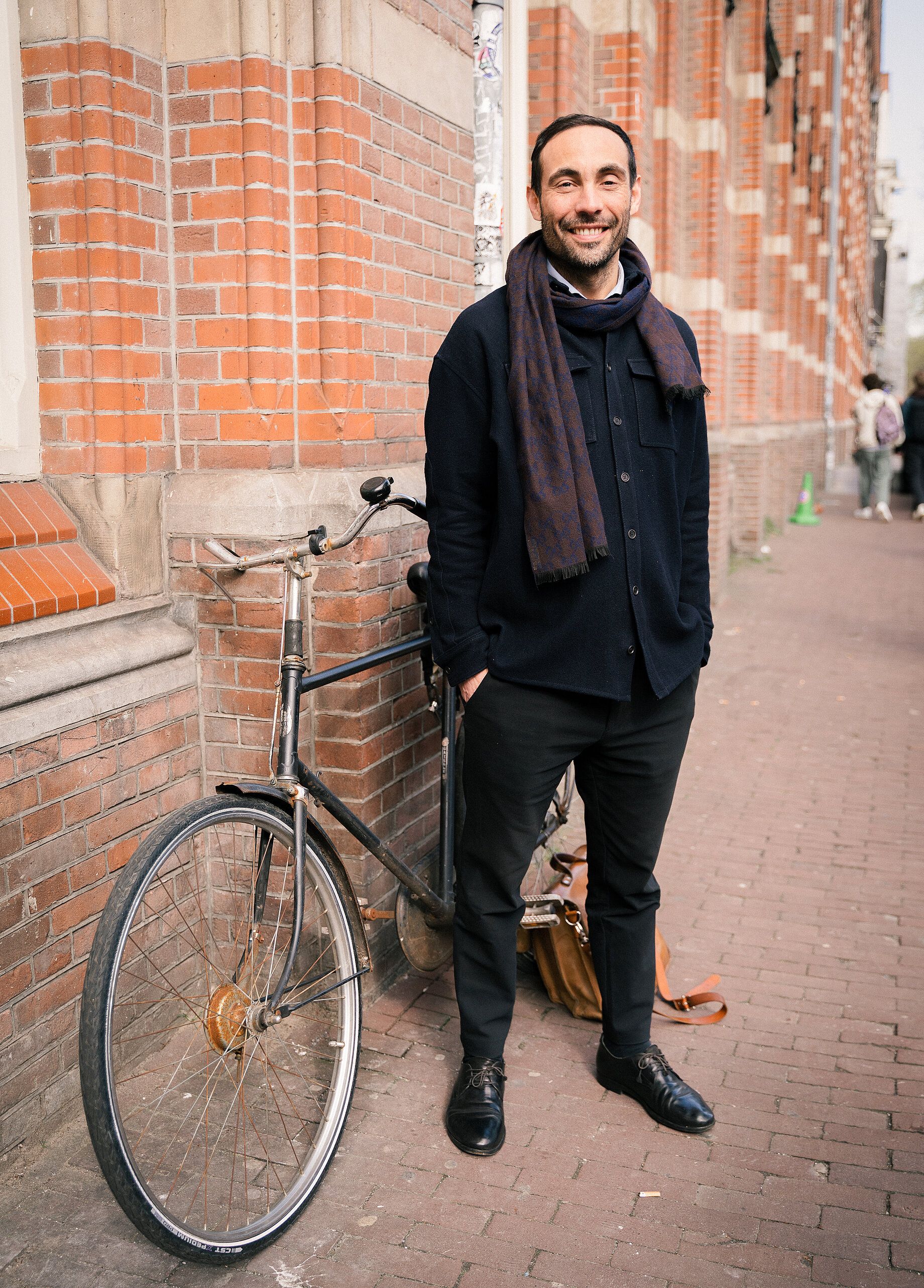 A man with short dark hair standing in front of a bike, who is wearing a dark jacket and a dark purple scarf
