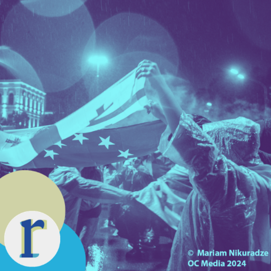 A person holding up a large flag with a blue field and gold stars during a rainy nighttime protest, with streetlights and a lit building in the background, in a blue duotone tint (photo credit: Mariam Nikuradze / OC Media, 2024).