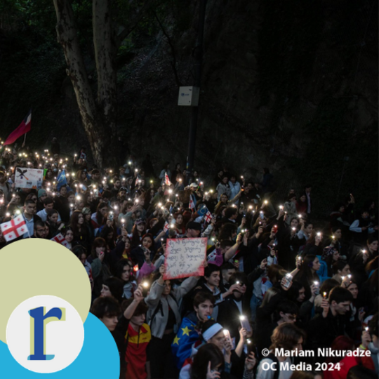 A large nighttime crowd holding up illuminated phone lights on a tree-lined street, with signs and flags including the Georgian flag, part of a 2024 demonstration in Georgia (photo credit: Mariam Nikuradze / OC Media).
