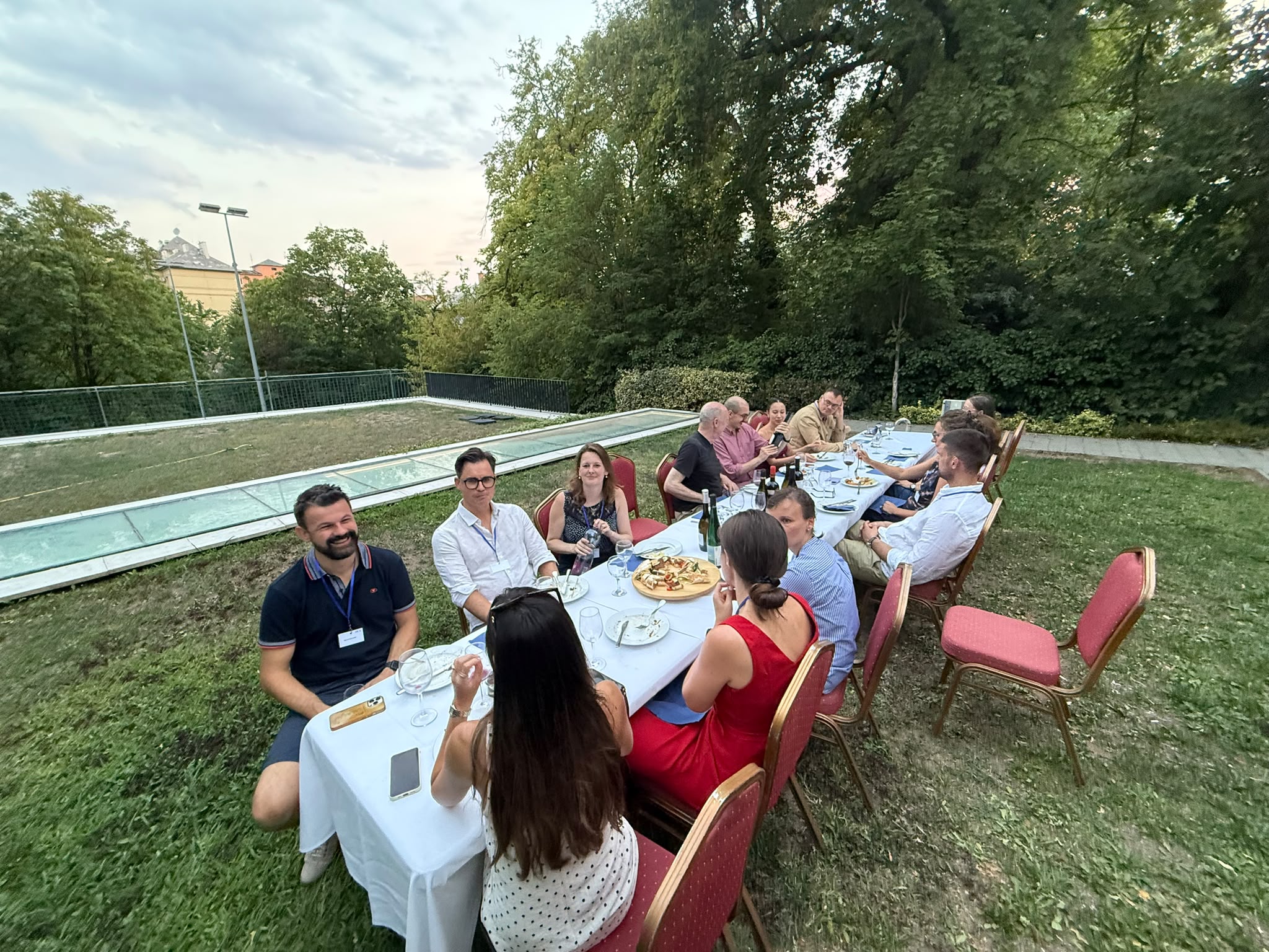 A dinner table is set up on lawn in a garden with ca. 15 participants of the summer university sitting around it, chatting.