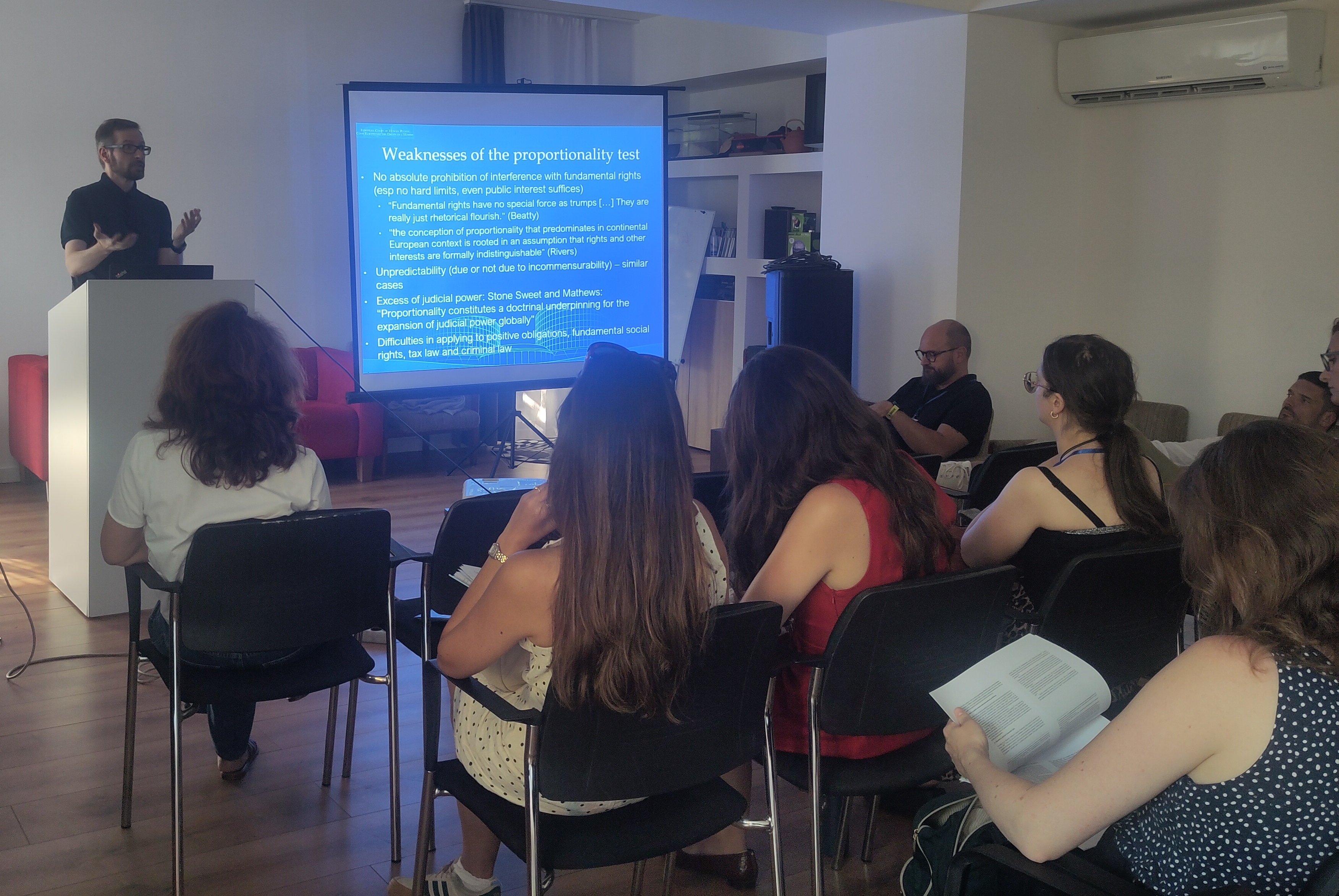 Photo taken from the back of a room, showing Andras Jakab speaking next to a powerpoint slide titled "Weaknesses of the proportionality test" in front of an audience