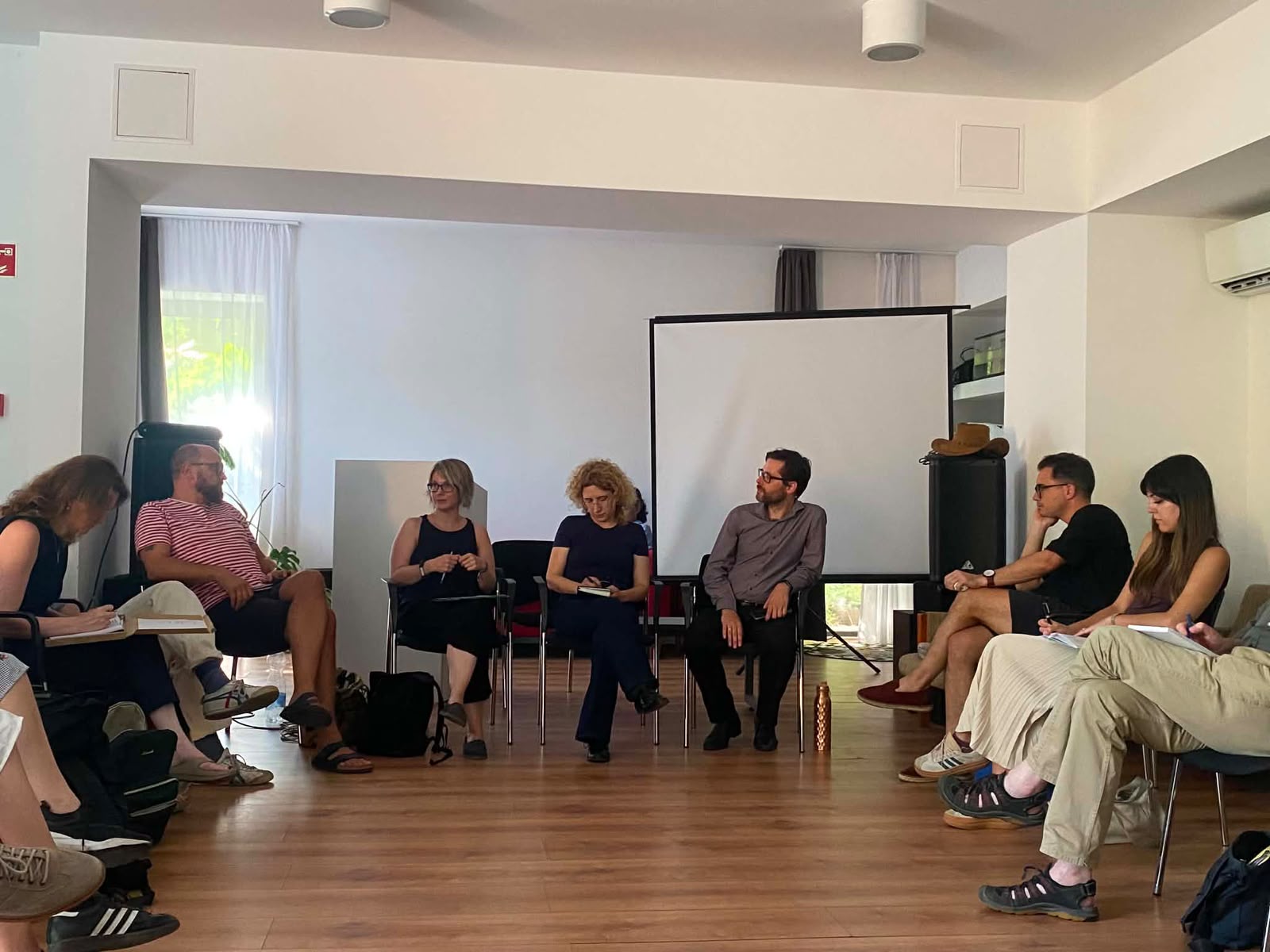 A group of adults sits in a circle in a bright room, appearing to participate in a panel discussion. The three panelists sit in a row at the front and several people are taking notes.